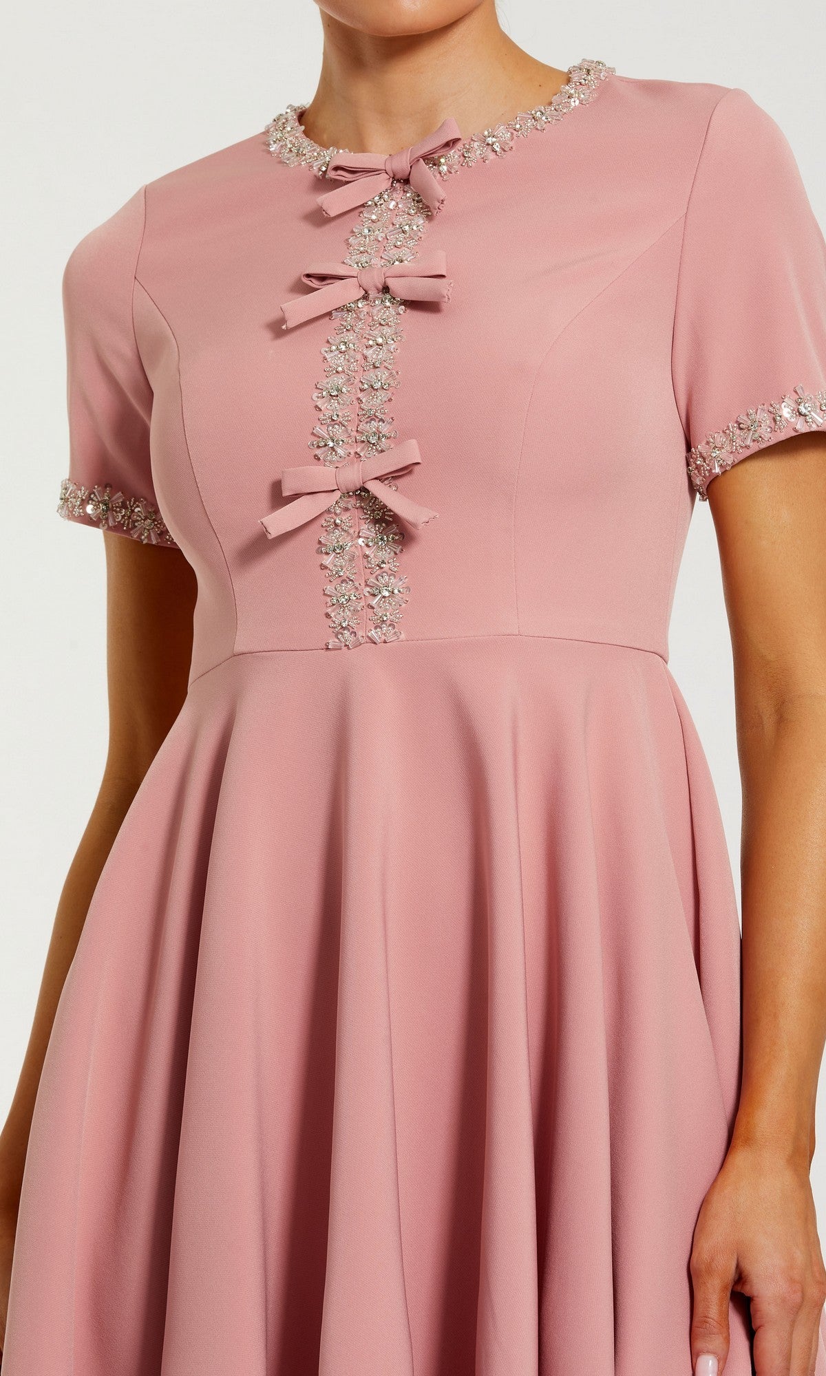 A woman wearing the Mac Duggal 56243 short party dressโa pink fit and flare mini with silver beaded trim accenting the sleeves, neckline, and three bows down the front.